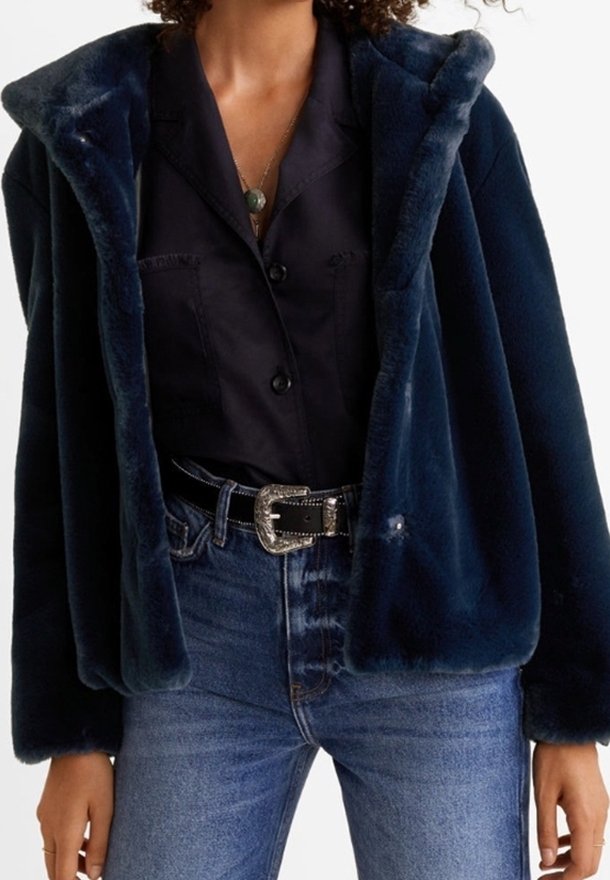 Femme portant une veste en fausse fourrure bleu marine par-dessus une chemise noire à boutons rentrée dans un jean bleu taille haute avec une ceinture à boucle argentée.