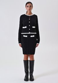 Vrouw in een zwarte gebreide cardigan en een bijpassende rok met witte bies en knopen, gecombineerd met kniehoge zwarte leren laarzen, rechtop staand.