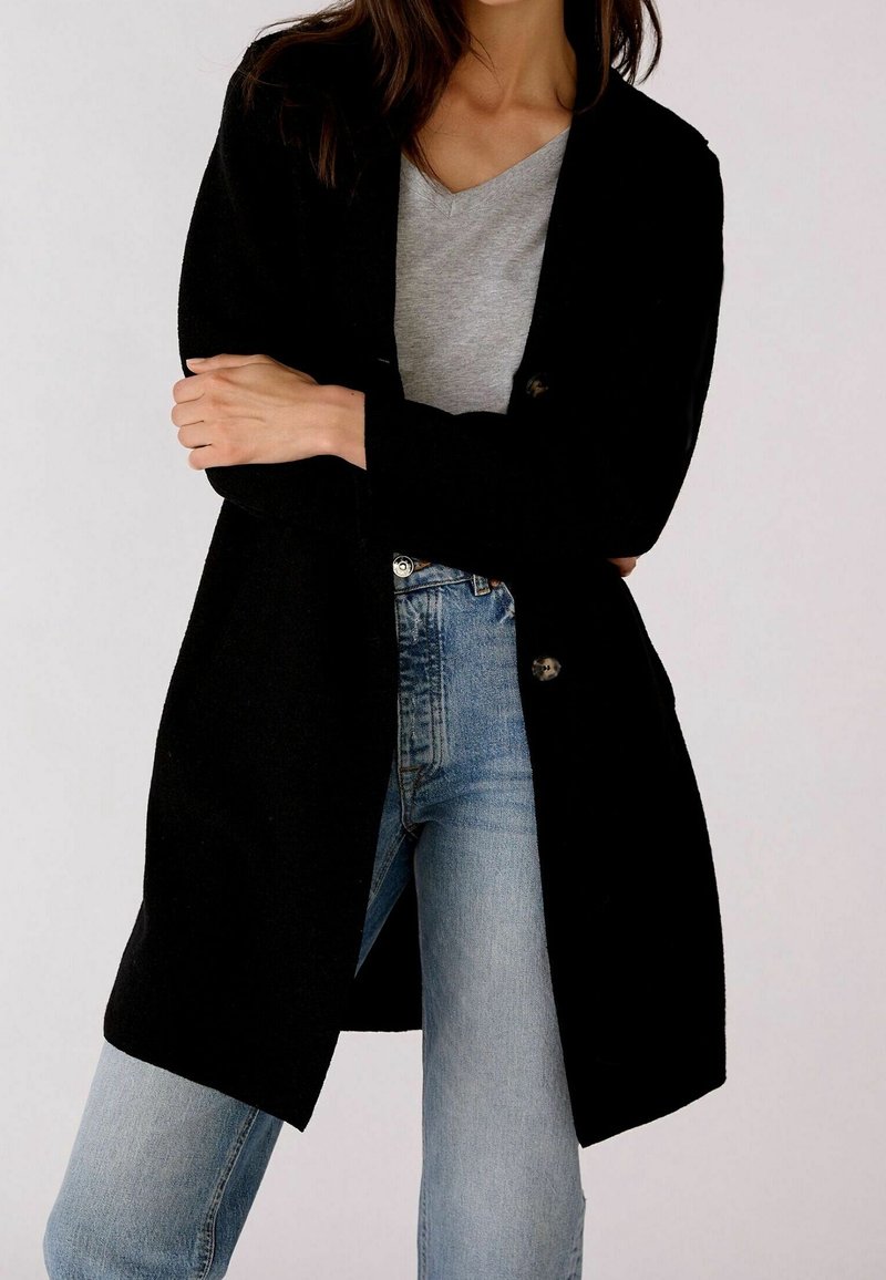Svart lång kofta med en slät yta, öppen frontdesign och två knappdetaljer, ihop med en grå V-ringad topp och blå jeans.