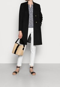 Abrigo largo negro con botones dobles, pantalones blancos, blusa con estampado, sandalias negras y un bolso de paja estructurado con acentos negros.