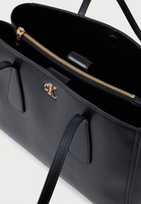 Μαύρη δερμάτινη τσάντα χειρός με φερμουάρ, χρυσά μεταλλικά στοιχεία και έντονο λογότυπο CK. Διαθέτει λεία υφή και διπλές ιμάντες ώμου.