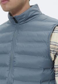 Veste isolante bleu clair avec un col haut, fermeture éclair et design matelassé. Surface texturée avec un rembourrage horizontal visible.