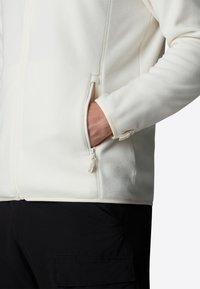 Chaqueta de forro polar crema con un bolsillo con cremallera, puños acanalados y un cuello alto. Textura suave, diseñada para calidez y comodidad.