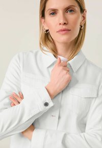 Weiße Denimjacke mit knöpfbarem Kragen, Brusttaschen und Knopfmanschettendetails. Glatte Textur und taillierte Passform.