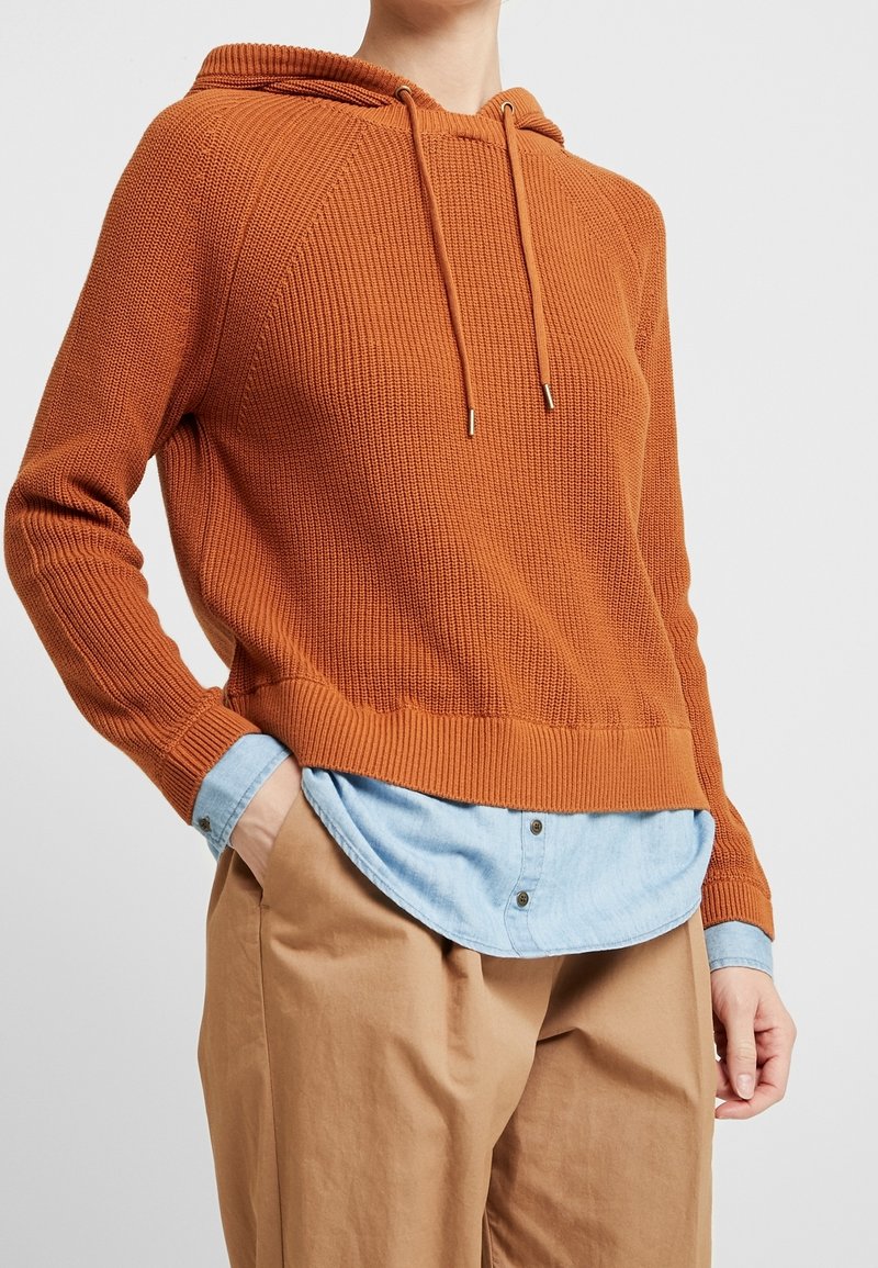 Ruosteenoranssi neulottu huppari, jossa on teksturoitu pinta, kiristysnauha ja lyhyt helma, kerrospukeutuneena vaaleansinisen napillisen paidan päälle.