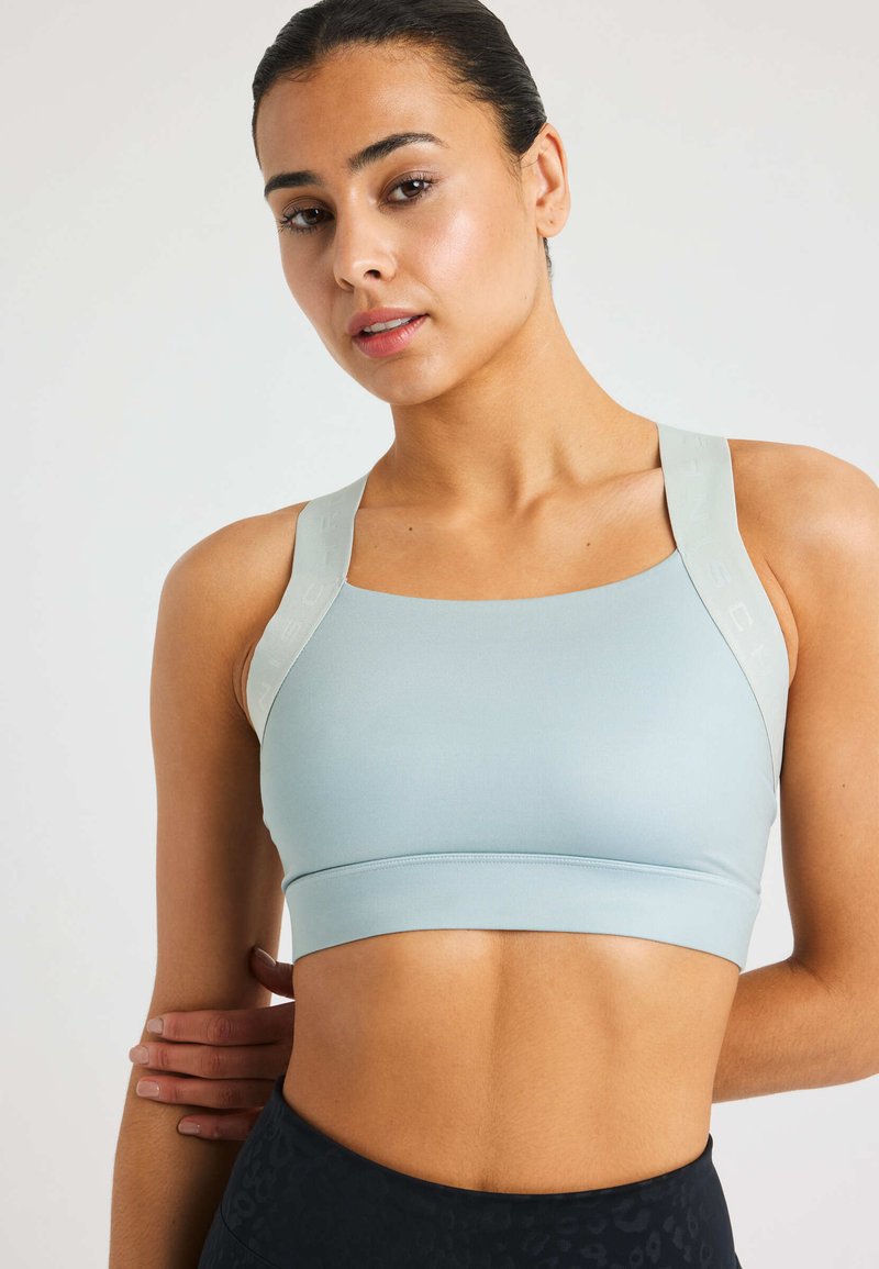 Vrouw met teruggebonden donker haar, gekleed in een lichtblauwe sportbh en zwarte high-waisted legging, staande tegen een effen achtergrond.