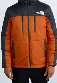 Mężczyzna ubrany w puchową kurtkę North Face w dwóch odcieniach czerwieni i czerni, z zasuwanymi kieszeniami na klatkę piersiową i z boku, stoi z rękami wzdłuż ciała.