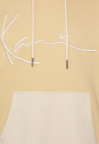 Ljusgul hoodie med vitt broderat text, med en ficka fram och dragsnören med silverdetaljer. Mjuk tygstruktur.