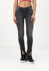 Jean gris foncé avec des fentes à l'avant au niveau des chevilles, fabriqué en tissu denim. Associé à des baskets noires avec des semelles blanches.