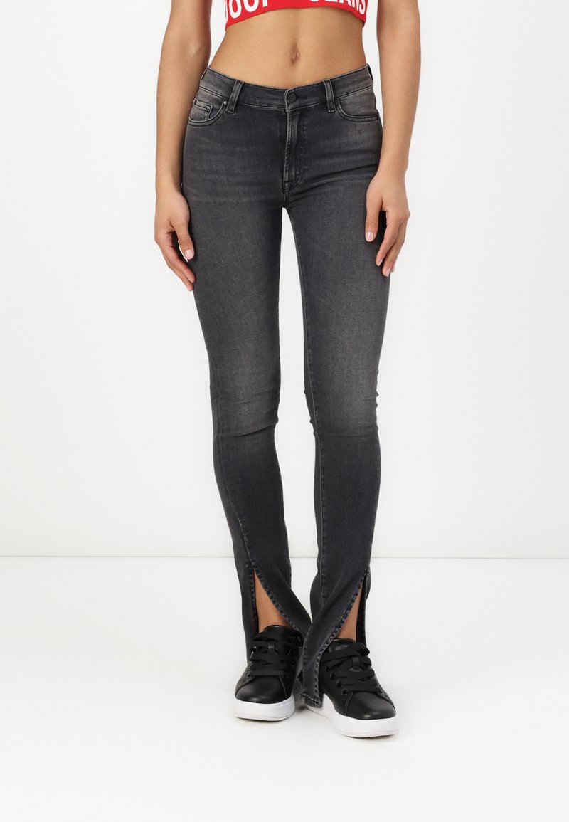 Jean gris foncé avec des fentes à l'avant au niveau des chevilles, fabriqué en tissu denim. Associé à des baskets noires avec des semelles blanches.