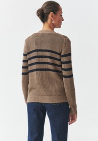 Knit sweater in tan with navy horizontal stripes, featuring a round neckline, ribbed hem, and long sleeves. Worn with dark blue jeans.