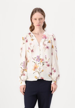 Kvinne iført en hvit blomstret langermet bluse med knapper og marineblå bukser, stående mot en enkel hvit bakgrunn.