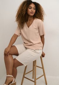 Ljusrosa kortärmad topp med V-ringning; tillsammans med beiga texturerade shorts. Modell sitter på en träpall mot en neutral bakgrund.