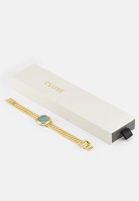 Gouden metalen horloge met een turkooise wijzerplaat en schakelarmband. Gecombineerd met een witte doos met "CLUSE" in goud gedrukt.