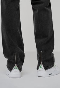 Pantaloni in denim nero con zip aperte alle caviglie, abbinati a sneakers bianche con un logo verde. La texture sembra morbida e consumata.