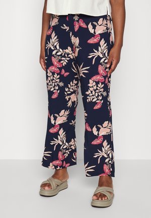 Pantalon large marine avec imprimé papillons roses et fleurs, porté avec des sandales beiges à brides croisées et un haut blanc.