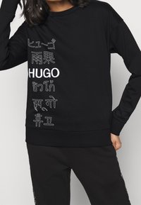 Persona che indossa un felpa nera con "HUGO" e il nome del marchio stampato verticalmente in vari caratteri asiatici sul lato sinistro frontale.