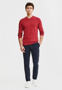 Czerwony sweter z długim rękawem i dekoltem w serek, zestawiony z ciemnogranatowymi spodniami i białymi trampkami. Slim fit, miękka tekstura, prosty design i brak widocznych wzorów.