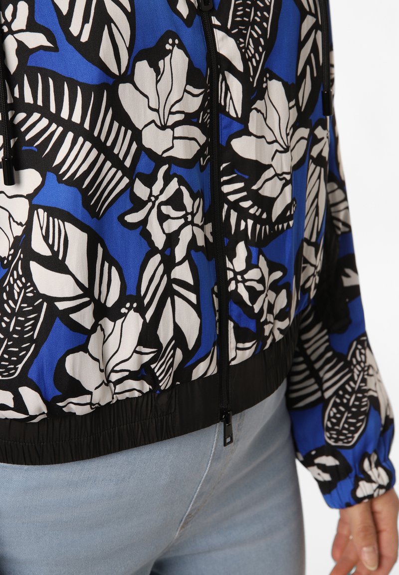 Blaue Jacke mit schwarz-weißen floral- und blattähnlichen Mustern, schwarzer Reißverschluss und elastischem Saum, getragen mit hellblauen Jeans.
