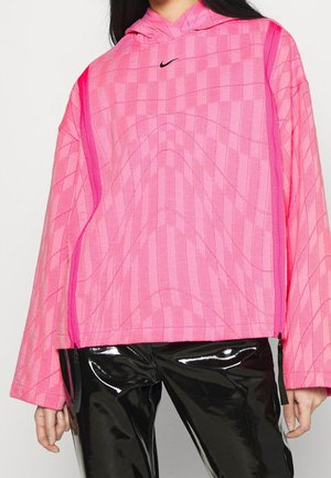 Kobieta w jaskraworóżowej, wzorzystej bluzie z kapturem i przednimi zamkami oraz czarnych, błyszczących winylowych spodniach, stojąca na jednolitym tle.