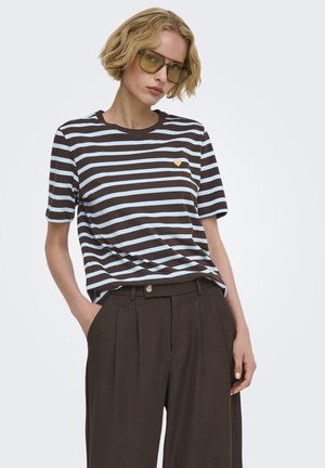 Femme aux cheveux blonds courts portant des lunettes teintées jaunes, un t-shirt rayé noir et bleu clair avec un petit cœur jaune, et un pantalon plissé foncé.