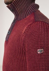 Strickpullover in Dunkelrot mit strukturiertem Körper, geripptem Hochausschnitt und drei Knöpfen; verfügt über ein Patch mit der Nummer "973."