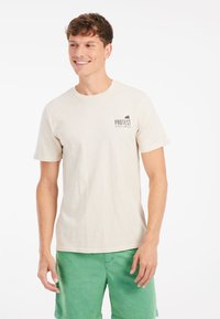 Beige bomulds T-shirt med korteærmer og rund hals; har et lille "PROTEST" logo på venstre bryst; båret med grønne shorts.
