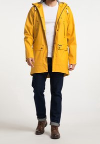 Man som bär en gul regnjacka över en vit skjorta, mörka jeans uppvikta vid fållarna och bruna snörskor, står mot en vit bakgrund.