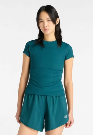 Ung kvinde med sort, glat hår iført aqua kortærmet skjorte og matchende sportsshorts, der står mod en hvid baggrund.