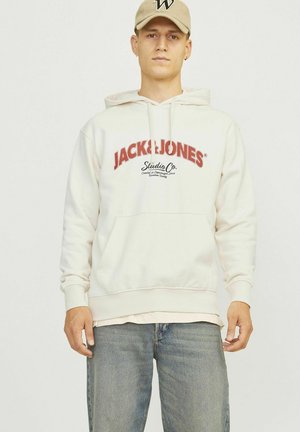 Weißer Kapuzenpullover mit rotem "JACK & JONES"-Logo und "Studio Co."-Text. Kängurutasche, gerippte Bündchen und Saum. Über hellblauen Jeans getragen.