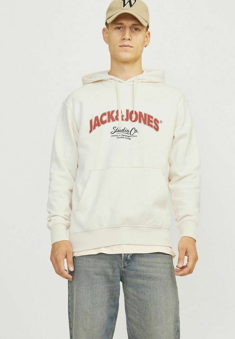 Sudadera blanca con capucha y logo rojo "JACK & JONES" junto al texto "Studio Co." Bolsillo tipo canguro, puños y dobladillo de canalé. Lleva puesta encima de unos jeans azul claro.