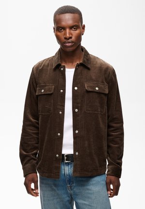 Man draagt een bruine corduroy button-up jas over een wit T-shirt en blauwe spijkerbroek, staand tegen een effen witte achtergrond.