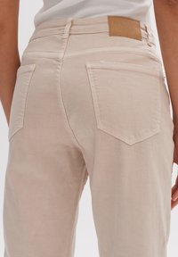Pantalon en denim beige à taille moyenne, avec poches plaquées à l'arrière et étiquette de ceinture en cuir marron clair. Le tissu présente une texture lisse et mate.