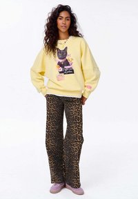 Žlutá veliká mikina s grafiku kočky a květinovými akcenty, zkombinovaná s hnědými kalhotami s leopardím potiskem a levandulovými botami.