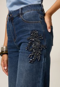 Jeans de denim en azul oscuro con intrincados bordados florales en cuentas negras en el muslo izquierdo. Presenta un diseño clásico de cinco bolsillos y una silueta ajustada.