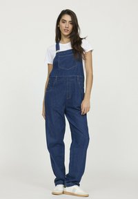 Salopette en denim avec un haut ajusté, des jambes larges et des poches avant. Dotée de bretelles réglables et de boutons en laiton sur les côtés.