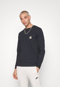 Hosszú ujjú, tengerészkék pamut ing, mellzsebbel és négyzet alakú logóval, fehér nadrággal viselve, amelyen fekete Nike logó található. Ezüst lánc nyaklánc.