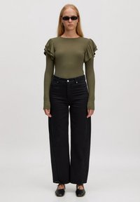 Olijfgroen geribbelde top met lange mouwen en ruches op de schouders, gecombineerd met hoge zwarte wijde jeans en zwarte platte schoenen.