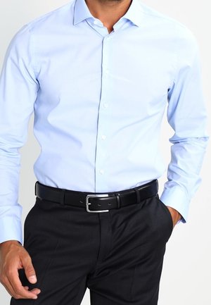 Mężczyzna noszący jasnoniebieską koszulę na guziki włożoną do czarnych spodni z czarnym paskiem, jedna ręka w kieszeni, na białym tle.