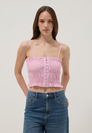Jeune femme portant un crop top froncé rose avec des boutons et des bretelles fines, associé à un jean en denim bleu taille haute.