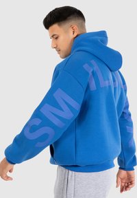 Haut à capuche bleu en tissu doux, avec un grand texte imprimé sur les manches. Le design comprend une capuche spacieuse et un fit décontracté.