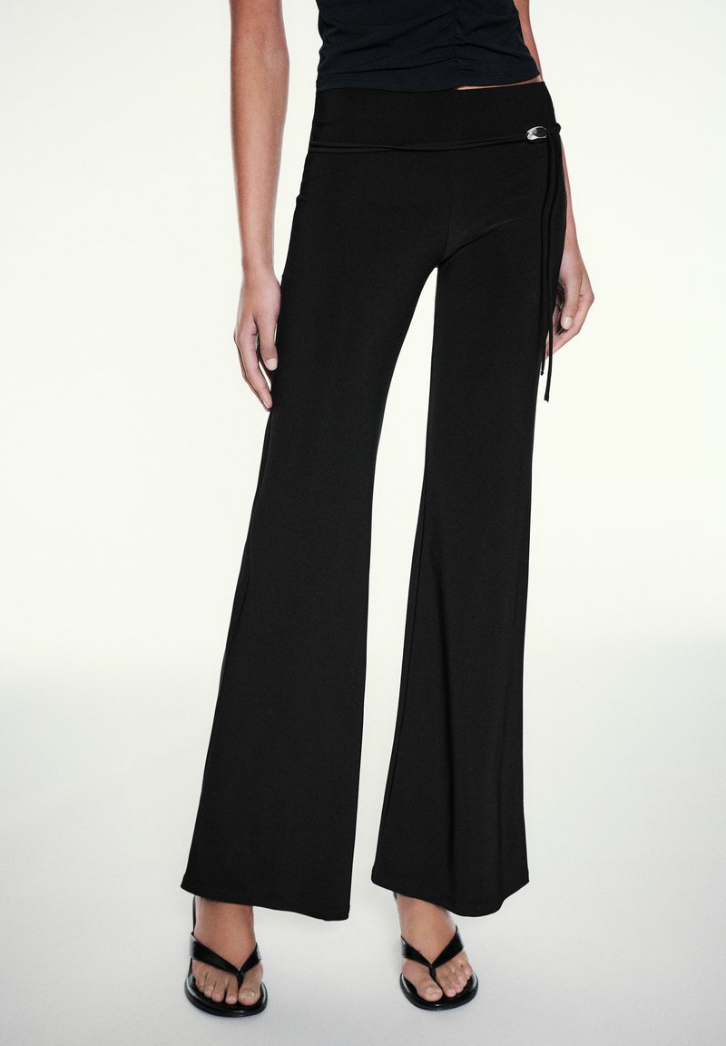 Femme portant un pantalon noir taille haute à jambes larges avec détail boucle argentée, associé à des sandales noires à lanières et un top noir plissé.