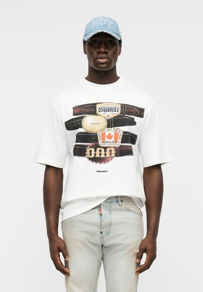 Άνδρας που φοράει λευκό γραφικό T-shirt με σχέδια ζώνης και λογότυπα, ανοιχτόχρωμο τζιν παντελόνι και ανοιχτό μπλε καπέλο με φθαρμένο κείμενο.