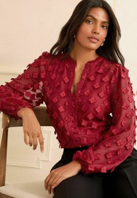 Blusa rossa trasparente con ricamo floreale, chiusura con bottoni sul davanti e maniche arricciate. Abbinata a pantaloni neri, evidenziando i dettagli del tessuto.
