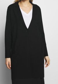 Svart lång cardigan med V-ringning, enkel knappstängning, långa ärmar och sidfickor. Mjuk textur och formell design.