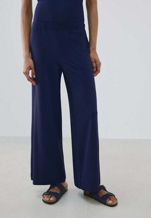 Persoon die marineblauwe wijde broek en marineblauwe sandalen met gespen draagt, staand op een lichtgrijze vloer tegen een effen achtergrond.