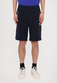 Pantaloni sportivi blu navy con cintura regolabile, tasche laterali e cuciture a contrasto. Indossati con scarpe da ginnastica bianche e calze nere.
