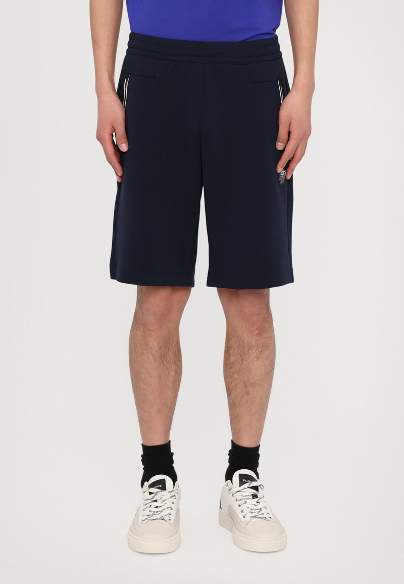 Pantaloni sportivi blu navy con cintura regolabile, tasche laterali e cuciture a contrasto. Indossati con scarpe da ginnastica bianche e calze nere.