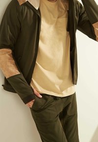 Chaqueta verde oliva con detalles en ante marrón claro en las mangas, combinada con una camiseta de color beige claro. El tejido parece suave, con un corte relajado.