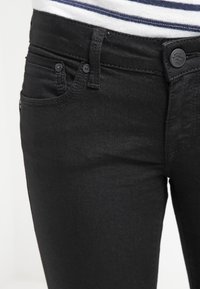 Černé džíny z denimu s přiléhavým střihem, vybavené zapínáním na knoflík, předními kapsami a jemnými švy. Část pruhované košile je viditelná nahoře.